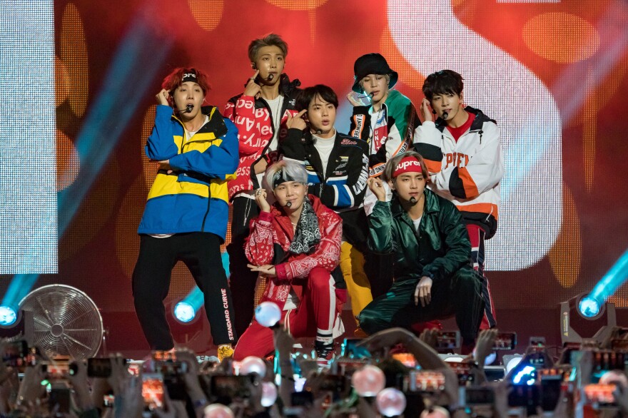 A K-pop group of seven young men pose on stage while performing in front of an audience, most of whom are holding mobile phones in the air to capture the moment.