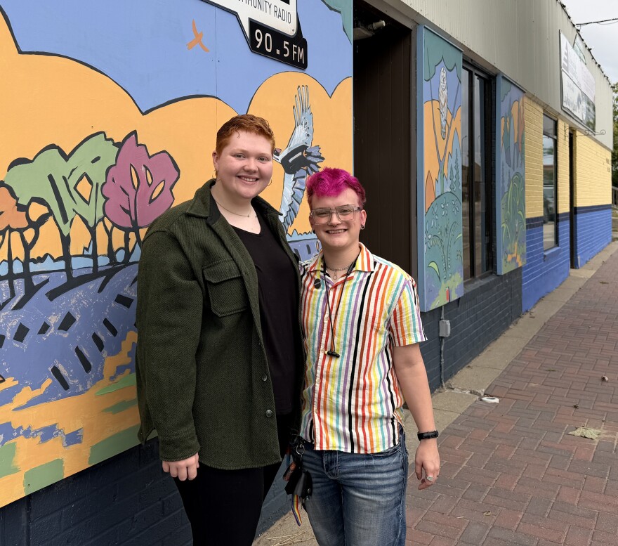 Two people wearing a dark green coat and various colored stripe shirt standing in front of a mural of nature elements.