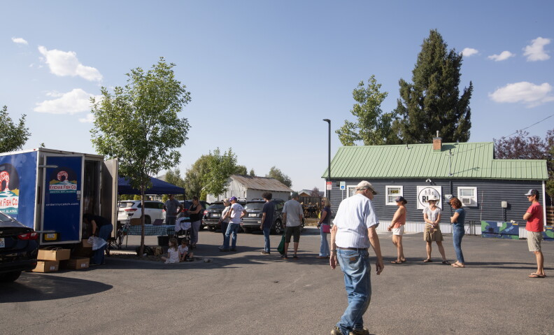 A line of people stand in a parking lot - one is walking in the foreground - waiting to get up to a blue tent and trailer. You can see a building in the background that says “Rise.”