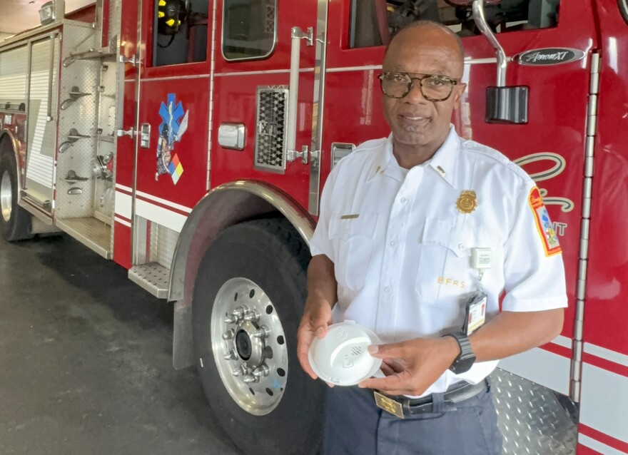 A uniformed fire department officer stands in front of a red fire truck holding a circular, white smoke alarm.
