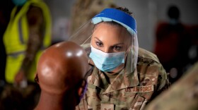 Female service member in face mask and shield speaks to a man sitting down.