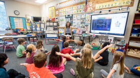 Elementary students are seated on the floor of a classroom facing their teacher, and are flapping their arms like they are flying.