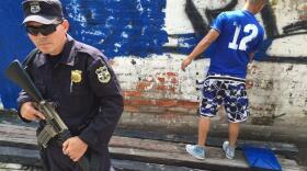 In Ciudad Delgado, a police officer guards a teen painting over gang tags. Conflicts between rival gangs are deadly, and hard for bystanders to avoid.