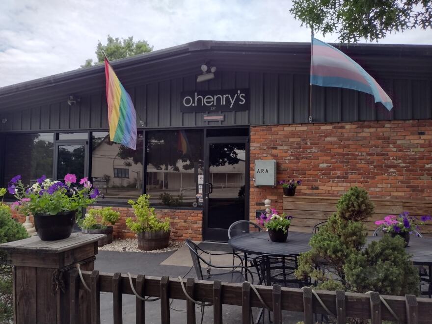 The brick front of O.Henry's with a pride flag, purple flowers, evergreens and patio seating. 