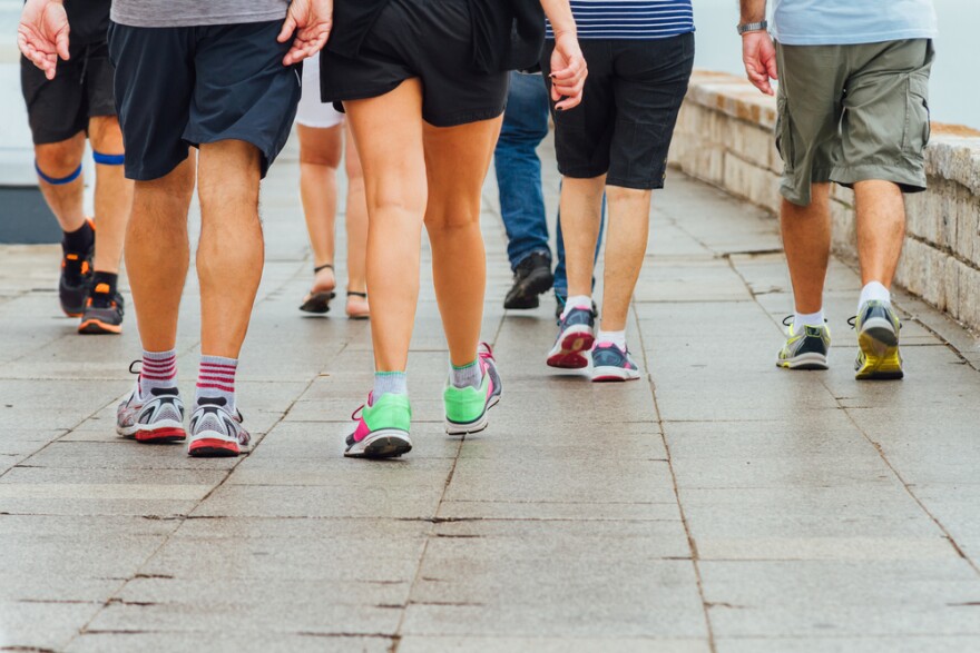 A group of people shown from the waist down in shorts and athletic shoes walking for exercise on a sidewalk.