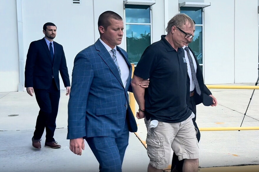 A man in a blue pin striped suit leads a man in handcuffs who is wearing a black t-shirt and cargo shorts off to the right. 