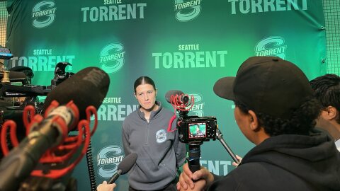 A woman in a gray crewneck sweatshirt faces reporters with microphones and cameras. The backdrop is green with the words and logo for the Seattle Torrent.