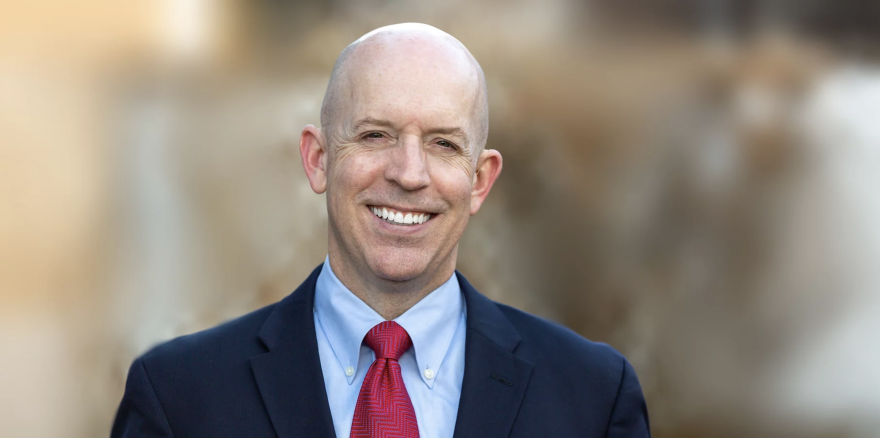 Adam Mayberry smiles for a headshot photo. He’s wearing a navy blue suit with a red tie. The background is out of focus.