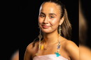 A woman poses against a black background. She is wearing a pink strapless dress and long beaded earrings. 