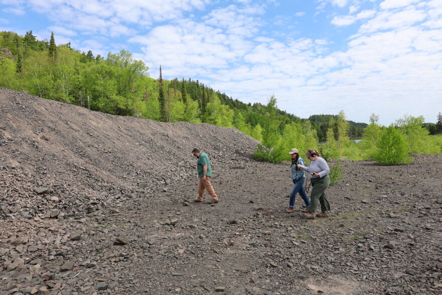 A white man in a green shirt and brown pants and two white women walk across a rock pile that sits at the bottom of the Greenstone Flow in the Keweenaw Peninsula. 