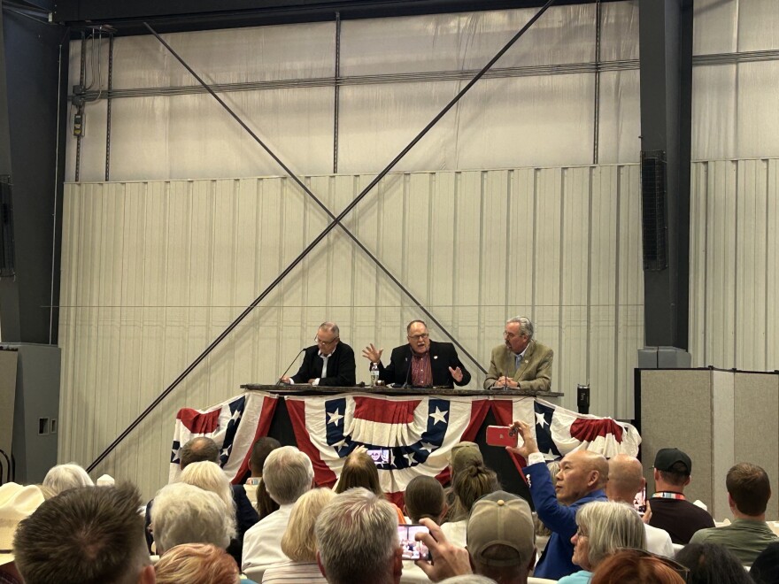 Three men sit at a table in front of a crowd