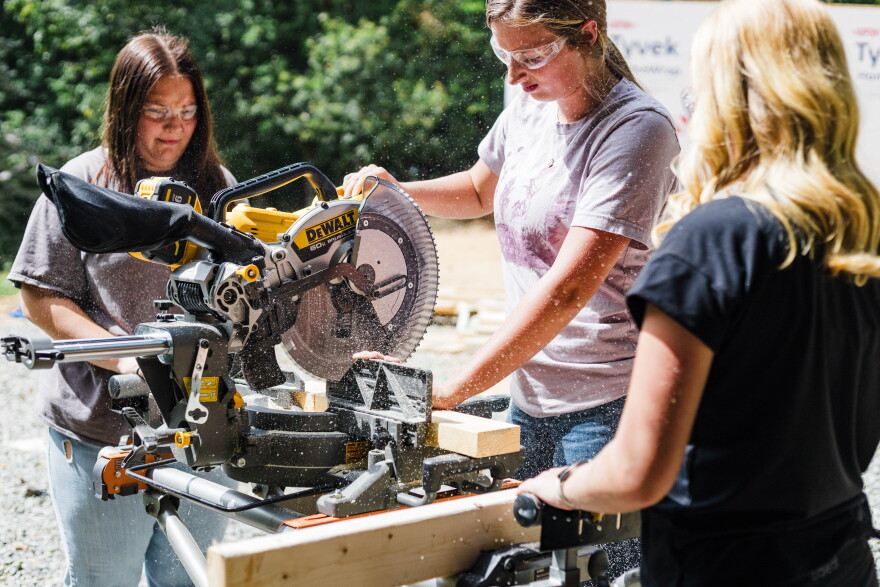 A young woman with a ponytail and clear work goggles uses a saw, with sawdust flecks flying in the air around her, as two other women stand watching. 