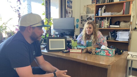A woman with long hair smiles across the desk at a man seated on the other side in a ballcap.