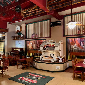 A restaurant dining room with several wooden tables and chairs positioned around a vintage organ.