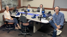 Four smiling people wearing headphones sit around a table in a radio talk studio: a woman front left has short grey hair and is wearing glasses, a white blouse, dark pants, and tan shoes; a woman back left has long light brown hair and is wearing a grey sweater; a man back center has short dark hair and a dark mustache and beard and is wearing a white button-down shirt under a grey suit coat; a man at right has very short dark hair and is wearing glasses, a blue button-down shirt, jeans, and grey and blue sneakers