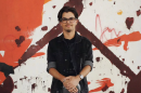 Man wearing glasses and casual clothes standing with hands clasped in front of him. Background is different paint colors splattered across a white canvas 