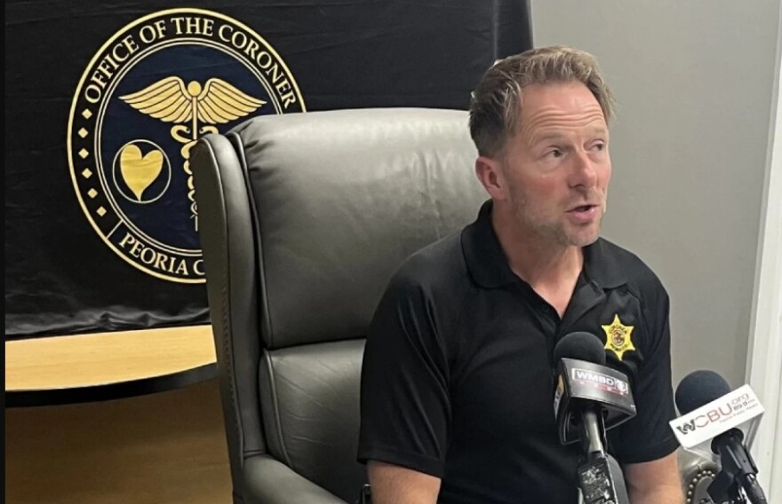 Peoria County Coroner Jamie Harwood speaks into media microphones with a banner behind his back at a 2024 news conference in his office.