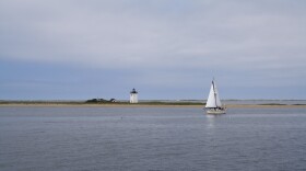 A Cape Cod rocky shoreline, with a lighthouse in the background and a small sailboat on the water