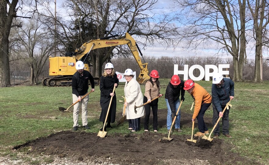 A group of men and women push gold shovels into a pile of dirt in a grass field, with trees, a construction vehicle, and large white letters spelling "hope" in the background.  