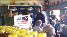 John James, a black man wearing a purple quarter-zip shirt, stands among a crowd of people who are clapping -- some are on their feet, and some are waving signs that say "James + Deboyer for Michigan". He is in a room with farmhouse-themed decorations, and there is a large banner behind him that reads "John James for Governor, Michigan". 