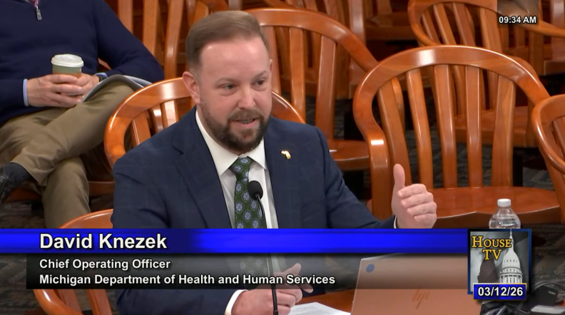 A man in a navy suit and green tie sits at a desk, talking and guesturing with his hand. There are wooden chairs behind him. Someone is sitting in one with his legs crossed and a disposable coffee cup in hand.
