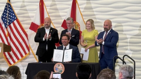 A man in a suit smiles and shows a document to a crowd. Three men in suits and a woman in a yellow dress stand behind him.