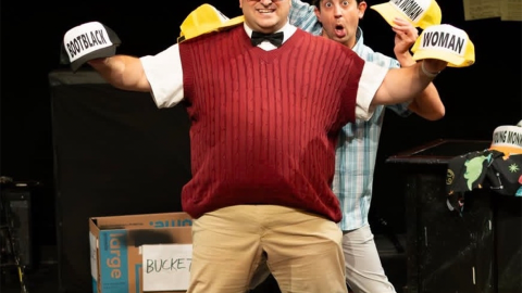 A man in a red sweater vest, a black bowtie, and khakis wears a red trucker hat that says "Monk" and holds two other trucker hats. Behind him, a man in a short sleeved plaid button up holds another trucker hat in one hand and a remote in the other. 
