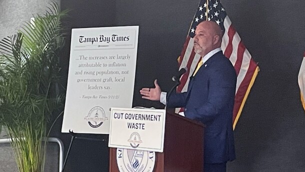 Florida CFO Blaise Ingoglia stands at a poduim that has a sign with the words "Cut Government Waste" hanging from it