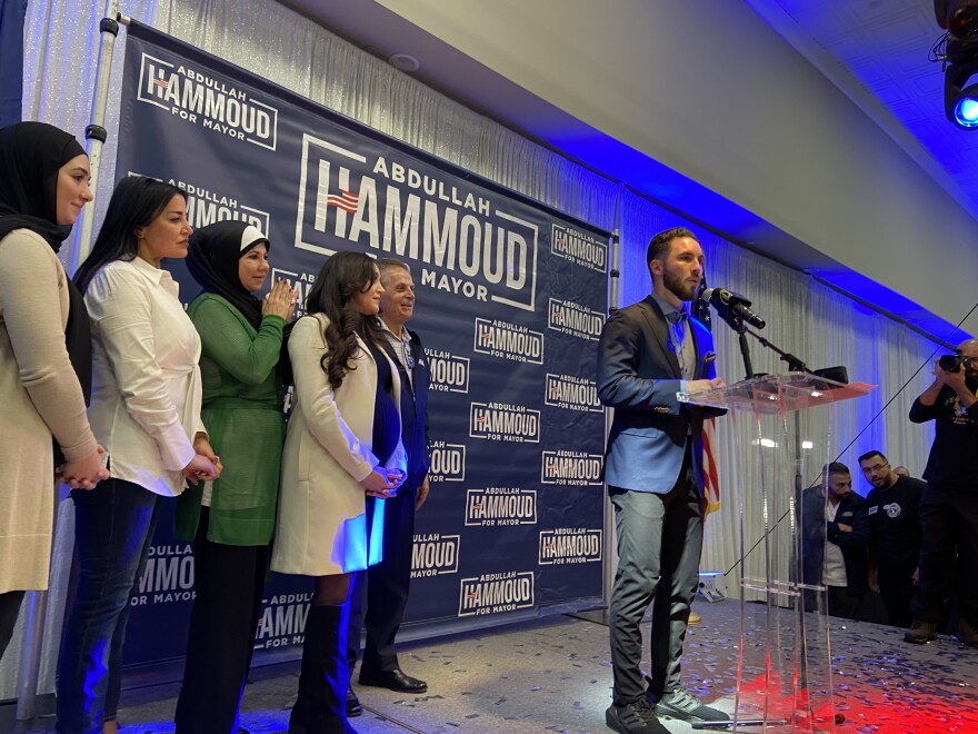 Abdullah Hammoud stands at a clear podium in front of a campaign sign. To his right stand four women and one man and to his left, off stage, are two men talking and one man with a camera 