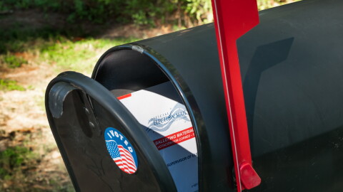 A black mailbox with an "I voted" sticker on it, slightly ajar. There is a mail-in ballot inside and the red mailbox flag is up.