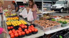 A woman in a t-shirt fills plastic bags with fresh produce as she stands behind tables of produce.