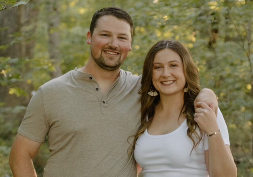 A man and a woman stand close together outdoors, smiling at the camera, with green trees and sunlight in the background. The man wears a light gray shirt; the woman wears a white shirt and earrings.