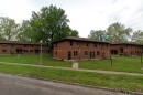 Three two-story red brick apartment buildings with brown roofs sit on a grassy lawn, surrounded by trees and sidewalks, under a cloudy sky.