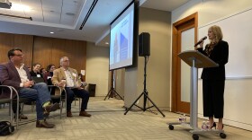 Mayor Mattie Parker, right, stands at a podium speaking into a microphone. The audience in the conference room listens attentively.