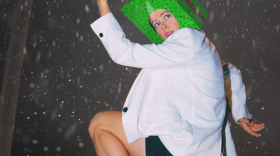 Holiday music, dancing Santas and present-box heads are all in store during Bare Bait Dance’s A Few of My Favorite Things holiday production. The photo shows a performer wearing a green gift box on their head, dressed in a white blazer and black boots, striking a dynamic dance pose amid falling snow. 