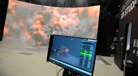 A computer screen in the foreground with a large immersive video screen behind it.