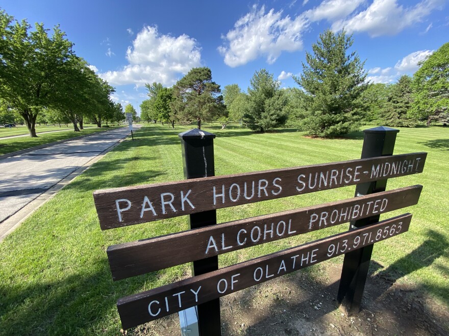 A wooden park sign inside a grassy park is surrounded by trees. The sign read "Park Hours Sunrise-Midnight, Alcohol Prohibited.