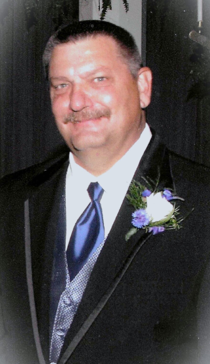 Man in a black tuxedo and purple vest with a purple tie and a white and purple carnation on the left lapel