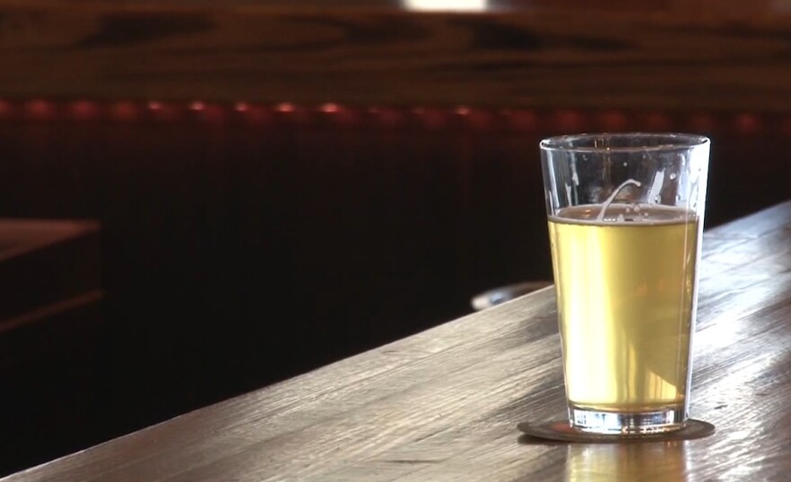 A pint glass of a light ale stands on a coaster on a wooden bar by itself.