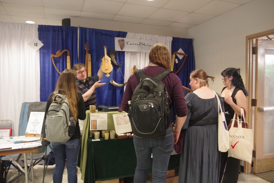 A vendor in a sweater vest and black shirt and khaki pants show off a wooden string instrument to four customers with backpacks and canvas tote  bags. His stall is in the exhibits hall for the 58th International Congress on Medieval Studies at Western Michigan University. Hanging on a blue curtained wall behind the vendor are small harps and horns, and other musical instruments popular in the Middle Ages.  