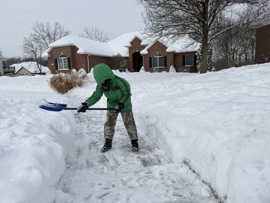 A child is surrounded by snow. He's wearing a bright green winter coat, camo snow pants, boots and gloves is shoveling snow off the sidewalk.