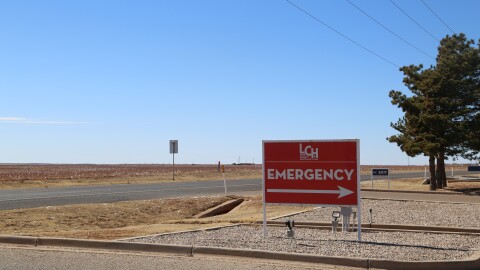 A photo of a red sign pointing to the emergency entrance to the Lynn County Healthcare System's hospital stands beside a long stretch of road and across from an empty field.
