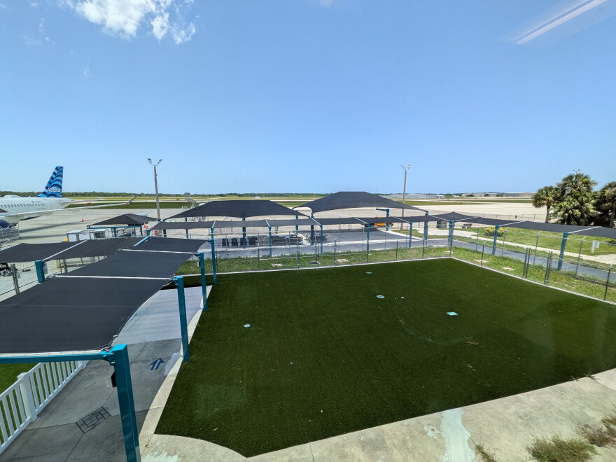 A row of shade canopies over an outdoor walkway leads to a fenced artificial-turf area at an airport terminal, with a JetBlue plane parked on the tarmac in the background under a clear blue sky.