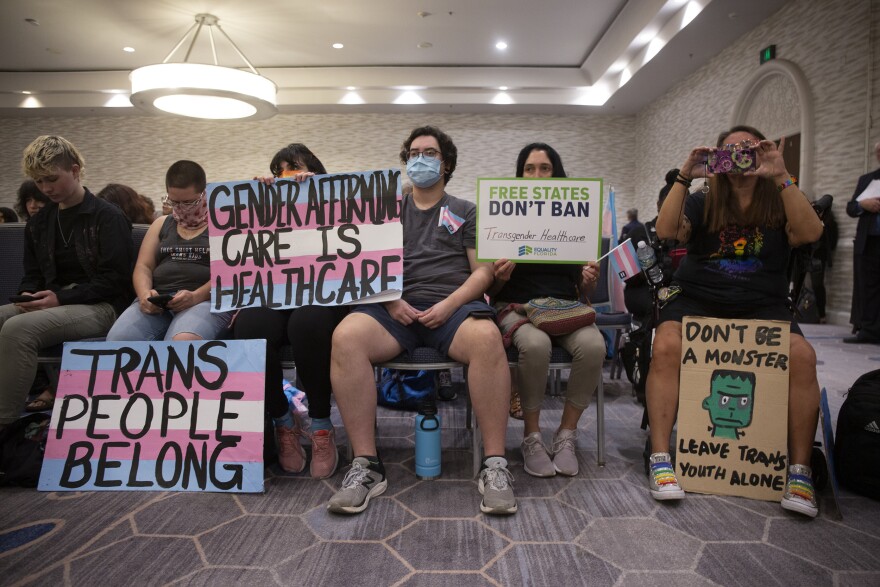 Group of people sit in chairs inside a meeting room. They hold signs painted with pink, blue and white, the trans pride colors. They read "trans people belong" and "Gender-affirming care is healthcare."