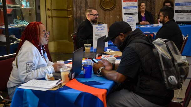 A woman in a light grey sweatshirt stis at a table with a blue tablecloth. A man in a black shirt sits across from her. In the background, two other pairs sit at different tables.