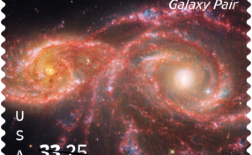 A detailed space image of two spiral galaxies interacting, with one galaxy curving around the other, displaying bright pink star-forming regions and swirling arms set against a black background filled with distant stars.