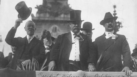 Historical black and white photograph from 1916 showing President Woodrow Wilson tipping his top hat alongside Indiana Governor Samuel M. Ralston and Indianapolis Mayor Joseph E. Bell as they view a parade from a reviewing stand in front of the Soldiers and Sailors Monument.