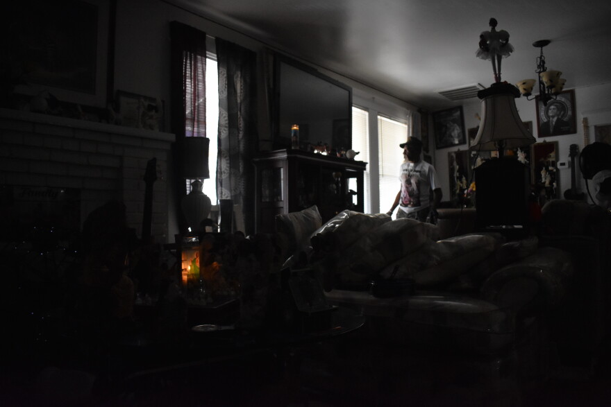  An older Black gentleman in a white T-shirt and an army hat stands by the window in his house that’s otherwise dark except for a couple candles. He said he is feeling crushed by his power bills from Tampa Electric Company