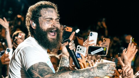 Man with curly brown hair and beard holding a microphone and surrounded by a crowd of people holding up cell phones