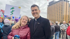 man in clerical collar and a woman in a raspberry-red jacket smile at camera